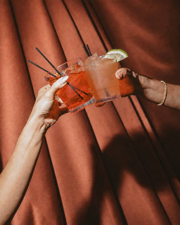Two people each holding colourful short cocktail glasses clinking them together in cheers, in front of a soft red floor-to-ceiling curtain.