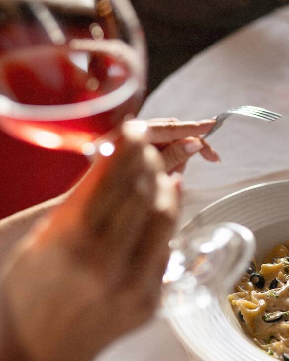 A closeup of a person moving their fork towards a beautifully plated pasta dish, artistically seen through an out-of-focus foreground wine glass with red wine.
